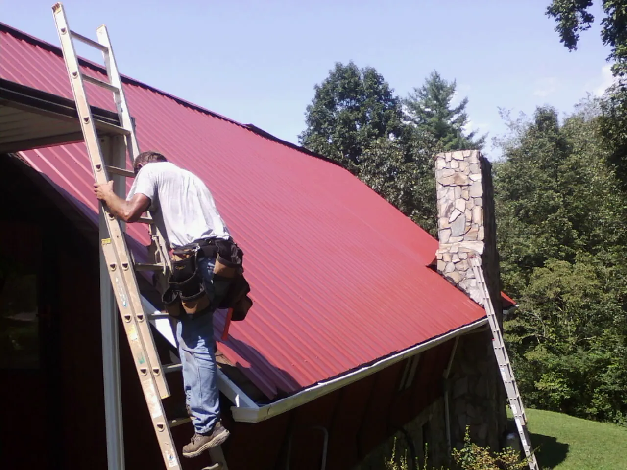 Roofer on a ladder beside a red metal roof and stone chimney — Soffit & Fascia Repair by Roofing Brothers in Maytown